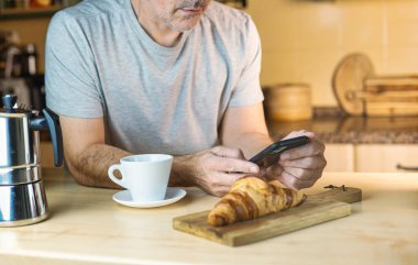 Kafkasyalı adam elinde kahve fincanı ve kruvasanla mutfakta cep telefonuna bakarak uyandı. Mutfakta sıcak ışık ve sıcak atmosfer var..