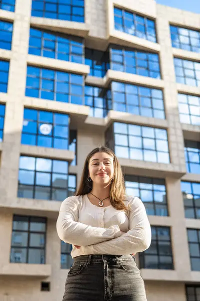 Beyaz bir kız cam bir binanın önünde duruyor, uzun saçlı, sakin, kendine güvenen bir ifade, kollarını göğsünde kavuşturmuş. Kendinden emin, iyimser tavır ve kameraya bakmak..