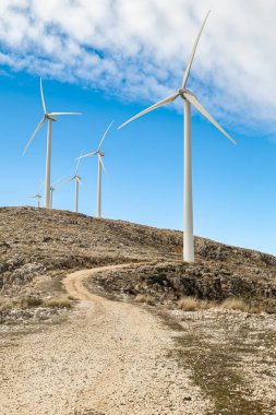 Ön planda, parçalı bulutlu bir gökyüzünün altındaki rüzgar türbininin dikey görüntüsü ve manzaranın içinden geçen bir yol. Doğada yenilenebilir enerji ve sürdürülebilirlik.