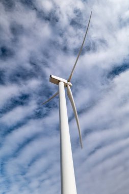 Bıçakları genişletilmiş, rüzgar türbini görünürde, mavi gökyüzü altında beyaz bulutlarla benekli, sürdürülebilir ve temiz enerjiyi ön plana çıkaran bir yel değirmeninin ayrıntılı görüntüsü..