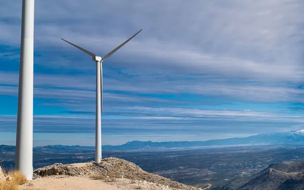 Dağdaki iki yel değirmeninin, bulutlu mavi bir gökyüzünün ve ovaların ve tepelerin arka planının yakın görüntüsü. Temiz ve alternatif enerji kavramı doğayla uyum içinde..