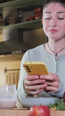Mutfakta çilekli smoothie içen ve seyyar telefona bakarken meyve yiyen genç bir kadın. Dikey formatta video, duyguları ve sağlıklı yaşam tarzını gösteriyor.