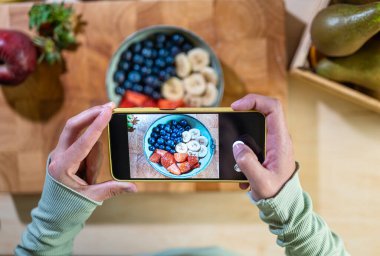 Kır mutfağında çilek, yaban mersini ve muzlu bir kase yakalarken cep telefonu tutan ellerin yakın çekimi. Gastronomik fotoğrafçılık sürecine odaklan.