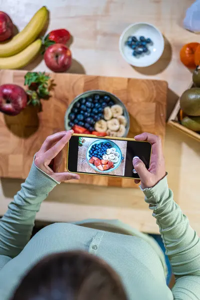 Cep telefonlu eller taze meyve kasesini çilek, yaban mersini ve muzla kırsal mutfakta yakalıyor. Yukarıdan gastronomik bileşim.