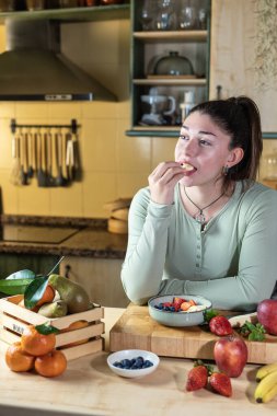 Mutfak masasına yaslanmış bir kadın, kâseden taze meyvenin tadını çıkarıyor. Doğal ışık ve rahat atmosferli dikey bir sahnede çeşitli meyvelerle çevrili..