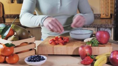 Bir kadının muz keserken orta boy kısa pozu, mutfakta çilek keserken. Elleri, kolları ve atölyenin bir kısmı, bir sepet meyve ve bir doğrama tahtasıyla görülüyor..