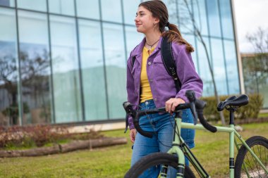 Bisikleti yanında yürüyen genç bir kadın. Sırt çantasını ve rahat ifadesini gösteren orta boy bir fotoğraf. Arka plan: Cam bina ve kentsel bitki örtüsü. Sürdürülebilirlik, bilinçli yaşam tarzı ve teknoloji ile bağlantı.