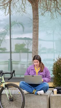 Genç bir kız dizüstü bilgisayarı üzerinde çalışırken elinde bir fincan karton kahve tutarak oturuyor. Park halindeki bisikleti ve arka planda yansımaları ve dekoratif bitkileri olan cam bir bina..