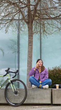 Genç bir kız beton bir bankta oturmuş cep telefonuyla sohbet ediyor. Onun yanında, park halindeki bisikleti ve arka planda yansımaları ve dekoratif bitkileri olan cam bir bina..