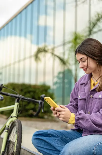 Genç kız oturuyor, cep telefonunu tutuyor ve konsantre bir ifadeyle sohbet ediyor. Bisikleti onun yanına park edilmiş. Sosyal ağ, dijital bağlantı ve kentsel yaşam tarzı.