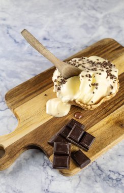 Bir fincan kısmen erimiş vanilyalı dondurmanın dikey görüntüsü, yanında çikolata parçacıkları ve yanında da çikolata parçacıkları. Hepsi tahta bir tahtanın üzerinde, ve fincanın içinde tahta bir kaşık var..