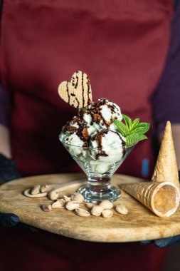 Ahşap bir tahtanın üzerinde servis edilen yeşil renkli dondurma, saçılmış şam fıstıkları, bir tutam nane ve kalp şeklinde bisküvi. Kırmızı önlük ve siyah eldiven giyen tanınmayan biri tarafından tutuluyor..