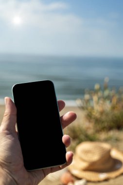 Akdeniz bitkileriyle dolu bir sahil. Ön planda, siyah ekranlı cep telefonunu tutan bir el. Arka planda, Akdeniz ve mavi gökyüzü.