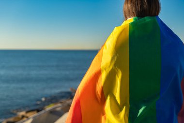 LGTBI bayrağına sarılı, yarı uzunlukta ve bir iskeleden denize bakan genç bir kadının yatay portresi. Doğal ışık, bayrağın yoğun renklerini aydınlatır ve vurgular.