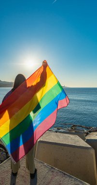 Denize bakan bir iskeleden gökkuşağı bayrağı kaldıran genç bir kızın arka ışığı. Arka planda mavi gökyüzü ve denizin olduğu açık bir ortamda özgürlük ve gurur. LGTBI, görünürlük, insan hakları
