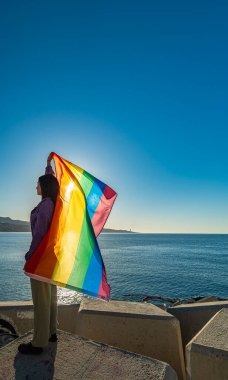 Profildeki bir kızın parlak bir görüntüsü. Rüzgâr onu açarken gökkuşağı bayrağını gururla kaldırıyor. Çeşitlilik, LGTBI görünürlüğü ve gençliğin doğal ortamdaki güçlendirilmesi.