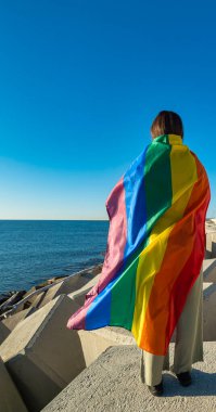 Sırtüstü yatıp denize bakan, LGTBI bayrağına sarılmış genç bir kadın. Bayrak modelin vücudunun çoğunu kaplıyor. Doğal ışık, bayrağın parlak renklerini ortaya çıkarır..