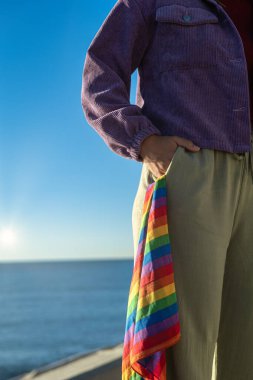 Omzundan dizine kadar uzanan dikey yarım figürlü görüntü. Bir el pantolonunun cebinde duruyor, ki bu da LGTBI gururunun sembolü olan gökkuşağı renginde bir eşarbın dikizlediği....