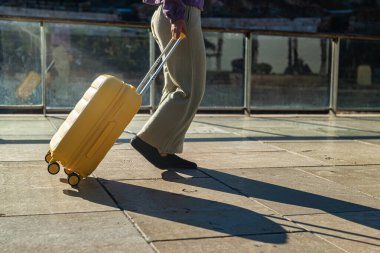 Malaga 'daki Roma tiyatrosunun çevresinde sarı bir bavulla şehir caddesinde yürüyen bir kadının bacaklarının yakın çekimi. Sabah ışığı uzun gölgeler yaratır.