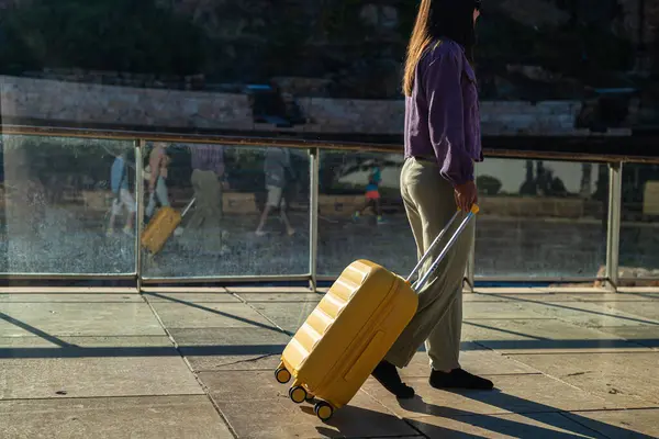 Roma tiyatrosunun önünde profilden yürüyen genç bir kadın, sarı bavullu, mor ceketli ve yeşil pantolonlu. Başkalarının bacaklarının yansıdığı yerde cam parmaklıklar var..