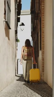 Sırtı dönük bir kız Malaga 'nın dar sokaklarında yürüyor, eski model deri sırt çantası ve sarı bavuluyla. Rahat ve otantik gezgin tarzı.