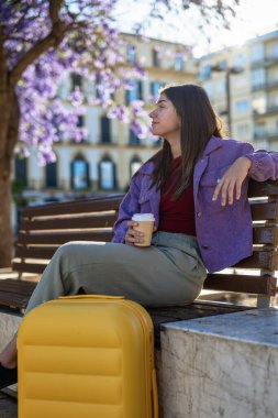 Sarı bavullu ve elinde kahveyle plaza bankında oturan genç bir kadın. Ilık arka ışık, gevşek ifade ve Malaga 'da sakin şehir ortamı.