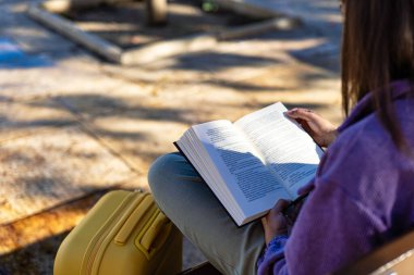 Bankta oturan bir kız yanında sarı bir valizle kitap okuyor. Malaga meydanında sakin bir sahne, yumuşak sabah ışığı ve rahat bir atmosfer..