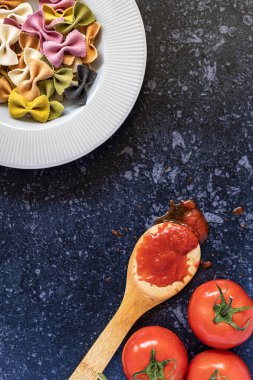 Dikey zenital fotoğraf. Sarmaşık domateslerinin yanında domates soslu kaşık. Üst: üç renkli farfalle tabağı. Beyaz arkaplan, sıçrama ve zanaatkar tarzı.