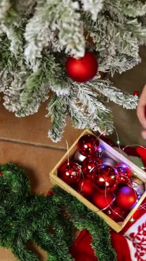 Yukarıdan dikey görüntü detayı. Noel süslemeleri ve elleri olan bir kutu. Ağaç dalına parlak bir top koyuyorlar. Bulanık arkaplan ve sıcak atmosfer.