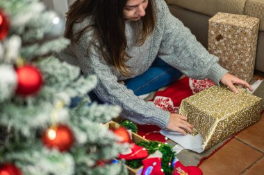 Yatay yakın çekim fotoğrafı. Bulanık Noel ağacı, yanında başka bir hediyeyle oturan kız..