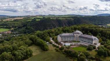 Aerial view of a large, elegant clinic hotel surrounded by lush greenery and rolling hills of Saalfeld, under a sky filled with fluffy clouds, offering a serene escape clipart