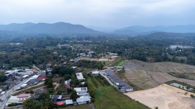 Tayland 'ın kuzeyinde, bulutlu bir gökyüzü altında tarlaların, evlerin ve uzak dağların yer aldığı kırsal bir manzarayı görüntüleyen hava manzarası doğal güzellik ve sükuneti gözler önüne seriyor.