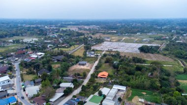 Geniş hava manzarası, Tayland 'ın kuzeyinde tarlaları, dağınık evleri ve bulutlu bir gökyüzünün altında dolambaçlı bir yolu olan bir kırsal araziyi gösteriyor.