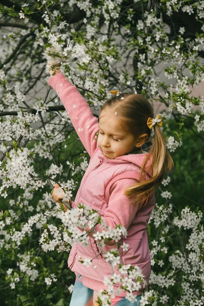 春の自然を探索する開花庭で素敵な小さな子供 木を揺らし花びらが頭の上に落ちてくる 庭の楽しみ — ストック写真