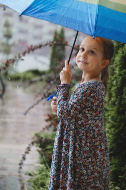 Şemsiyeli bir çocuk yağmurun altında yürüyor, gökkuşağı renkli şemsiyeli mutlu bir kız evin yanında, sıcak ve nemli bir yaz havası ve su birikintileri var. 