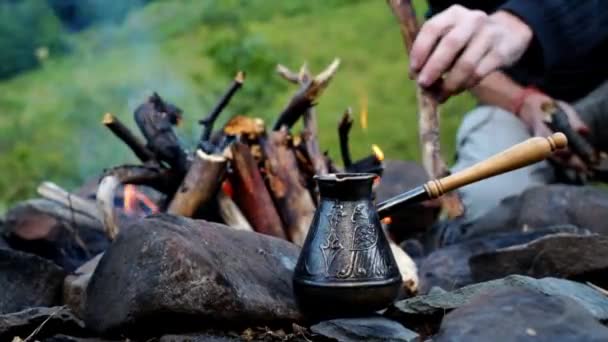 在山上旅行的时候 在篝火边煮咖啡 土耳其咖啡在户外篝火上的特写 男人的手把木头放进篝火里 徒步旅行时喝早茶 — 图库视频影像