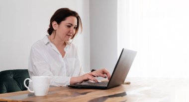 Orta yaşlı serbest çalışan bir kadın evde çalışıyor. 40 'lı yaşlarda oldukça büyük bir kadın sabahları mutfağında dizüstü bilgisayar ve telefonuyla bir bardak sıcak içecekle çalışıyor. Gerçek insan yaşam tarzı.