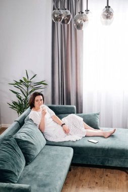 Sabahları sıcak ev atmosferinde kahve içen bir kadın. Orta yaşlı, beyaz elbiseli bir kadın kanepeye uzanmış, modern bir evde dinleniyor. Bir fincan çayla güneşli bir gün, kanepede uzanmak...