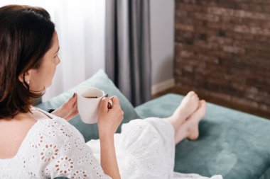 Sabahları sıcak ev atmosferinde kahve içen bir kadın. Orta yaşlı, beyaz elbiseli bir kadın kanepeye uzanmış, modern bir evde dinleniyor. Bir fincan çayla güneşli bir gün, kanepede uzanmak...