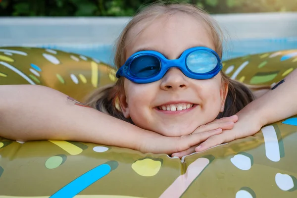 Happy preschool girl in outdoor swimming pool on inflatable ring. Child enjoying summer time swimming in backyard. Kid having fun floating in water, activities for rest and health. Funny face