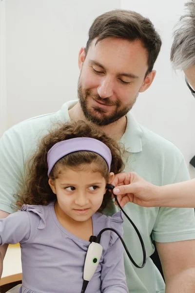 Child hearing test. Doctor audiologist examining kids ear and assess disorder of hearing using screening, diagnostic technology. Girl sitting on fathers laps in hospital or clinic