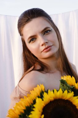 Beyaz kumaşlı arka planda ayçiçekleri olan romantik bir kadın. Temiz ten ve doğal güzellik. Kırsal yaz tatili