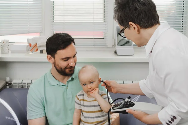 Çocuğun otolaringolojide işitme sınavı var. Küçük bir hastayı muayene etmek, çocuk işitme testini odiyologdan duymak. Babayla bebek doktoru muayene ediyorlar.