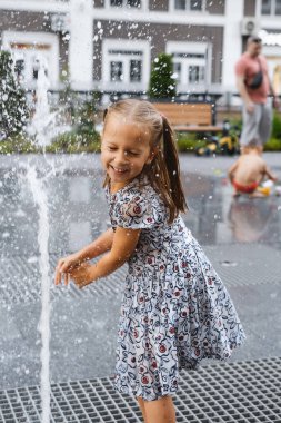 Şehirdeki çeşmede oynayan bir çocuk. Sıcak yaz havası. Su fıskiyeleriyle oynayan, koşan ve gülen mutlu kız. Aktif çocukluk. Açık havada oyunlar. Modern şehir sokağı