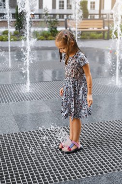 Şehirdeki çeşmede oynayan bir çocuk. Sıcak yaz havası. Su fıskiyeleriyle oynayan, koşan ve gülen mutlu kız. Aktif çocukluk. Açık havada oyunlar. Modern şehir sokağı