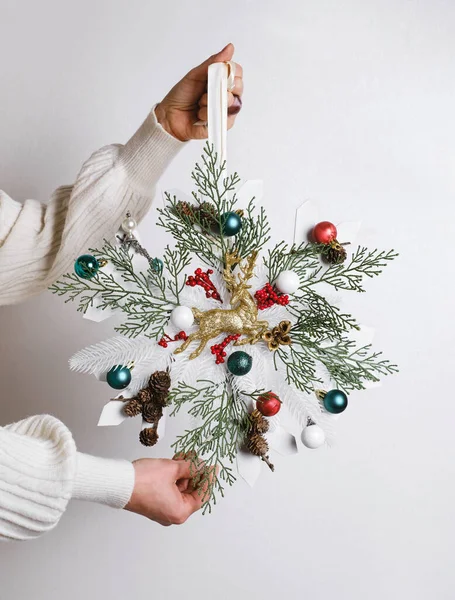 トナカイの白い杉の基盤および円錐が付いている雪片の形のクリスマス リースを握る閉鎖した女性 女性の手の無駄のお祝いの装飾無駄 グリーンブルーチとボブル ホワイトでクローズアップ — ストック写真