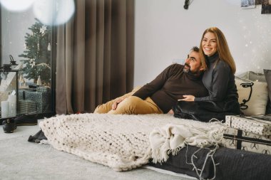 Romantik Noel arifesini evde kutlayan mutlu bir çift. Aile partisi için Noel iç dekorasyonu. Boşluğu kopyala, tebrik kartı. Orta yaşlı bir adam ve kadın örgü örmüş bir battaniyeyle rahat bir yatakta oturuyorlar.
