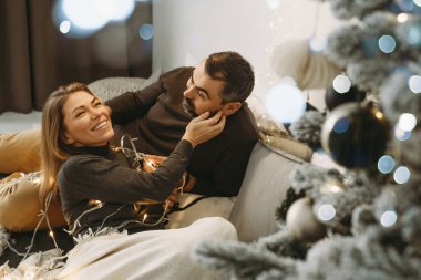 Romantik Noel arifesini evde kutlayan mutlu bir çift. Aile partisi için Noel iç dekorasyonu. Boşluğu kopyala, tebrik kartı. Orta yaşlı bir adam ve kadın örgü örmüş bir battaniyeyle rahat bir yatakta oturuyorlar.