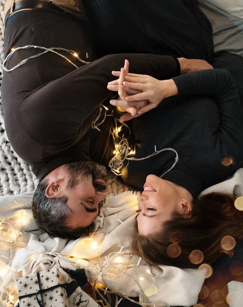Happy couple celebrating romantic Christmas eve at home. Christmas interior decoration for family party. Copy space, greeting card. Middle-aged man and woman sitting on cozy bed. Top view