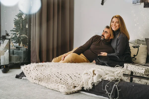 Happy couple celebrating romantic Christmas eve at home. Christmas interior decoration for family party. Copy space, greeting card. Middle-aged man and woman sitting on cozy bed with knitted blanket